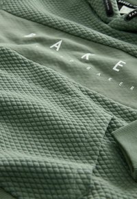 Sweat à capuche vert avec une texture matelassée, texte logo blanc et cordon de serrage. Le matériau semble doux et légèrement texturé.