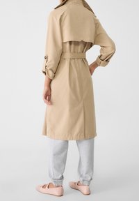 Trench-coat beige avec une taille ceinturée, une fente arrière ouverte et des manches boutonnées, accompagné d'un pantalon de survêtement gris et de ballerines rose pâle.