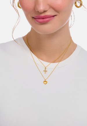 Goldfarbene, geschichtete Halsketten mit einem herzförmigen Anhänger und einem Kreuzanhänger an filigranen Ketten, getragen über einem weißen T-Shirt.