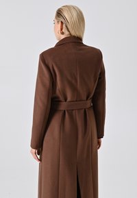Donna con capelli biondi corti, che indossa un lungo cappotto marrone con cintura, in piedi rivolta di spalle su uno sfondo chiaro e semplice.