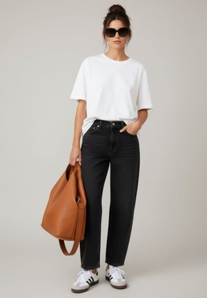 Vrouw draagt een oversized wit T-shirt, zwarte spijkerbroek, witte sneakers, zonnebril, houdt een grote bruine leren tas vast, staand tegen een effen achtergrond.