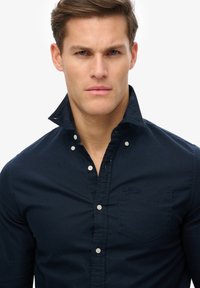Navy blue button-up shirt featuring a collar, long sleeves, and a chest pocket with a small brand logo. Cotton material with a smooth texture.