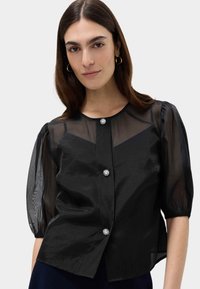 Zwarte blouse met doorschijnende puffmouwen, ronde halslijn en drie decoratieve bloemenknopen aan de voorkant. Glad stof met een lichte glans.