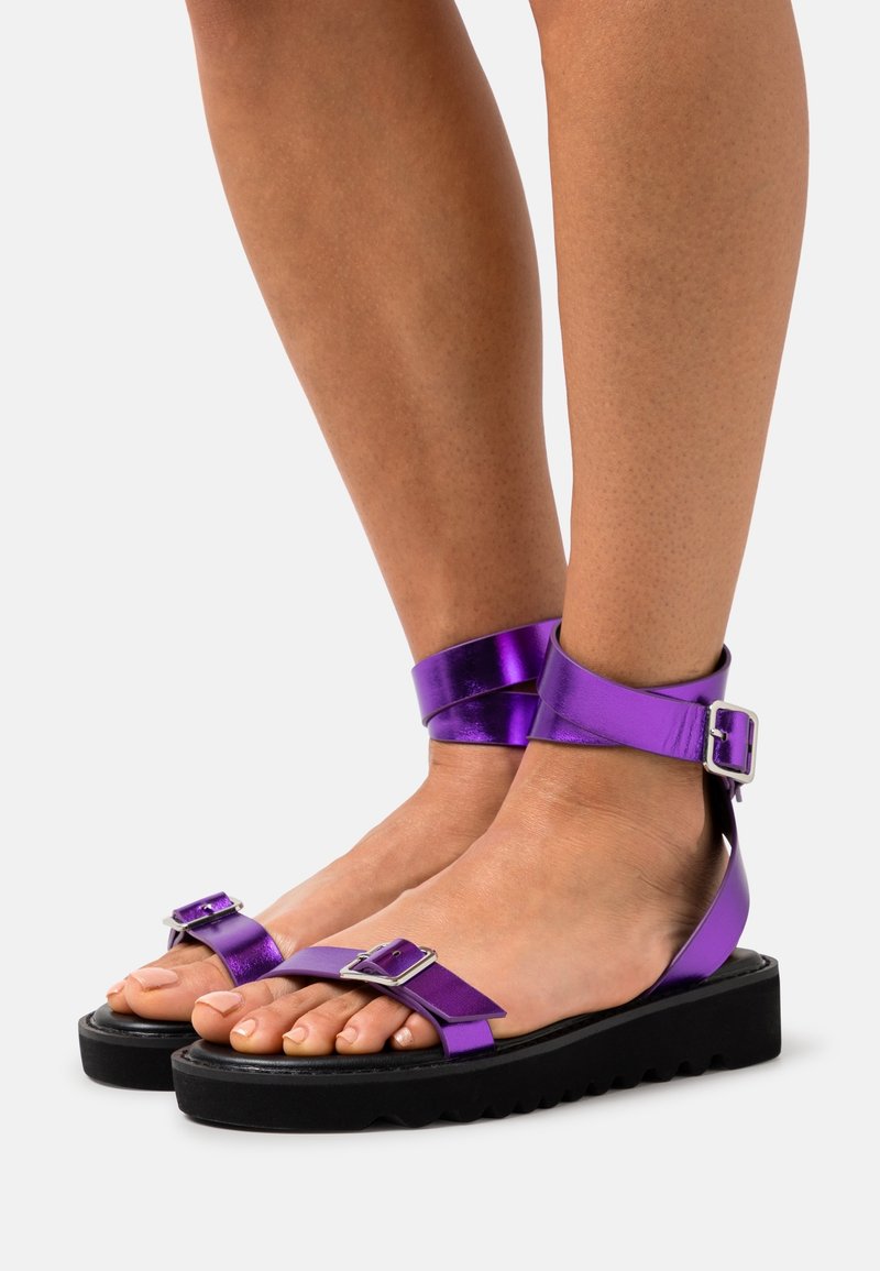 Pieds nus portant des sandales métalliques violettes avec des brides à la cheville et aux orteils et des semelles épaisses noires sur fond blanc.