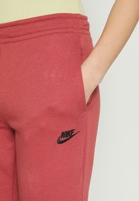 Person med röda Nike träningsbyxor, med handen i fickan, som visar den svarta Nike-logotypen på vänstra lår, mot en ljus bakgrund.