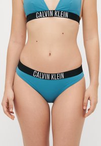 Top de bikini em triângulo turquesa com cintura preta com logotipo, apresentando uma textura suave e design minimalista. Letreiro da marca em branco na faixa.