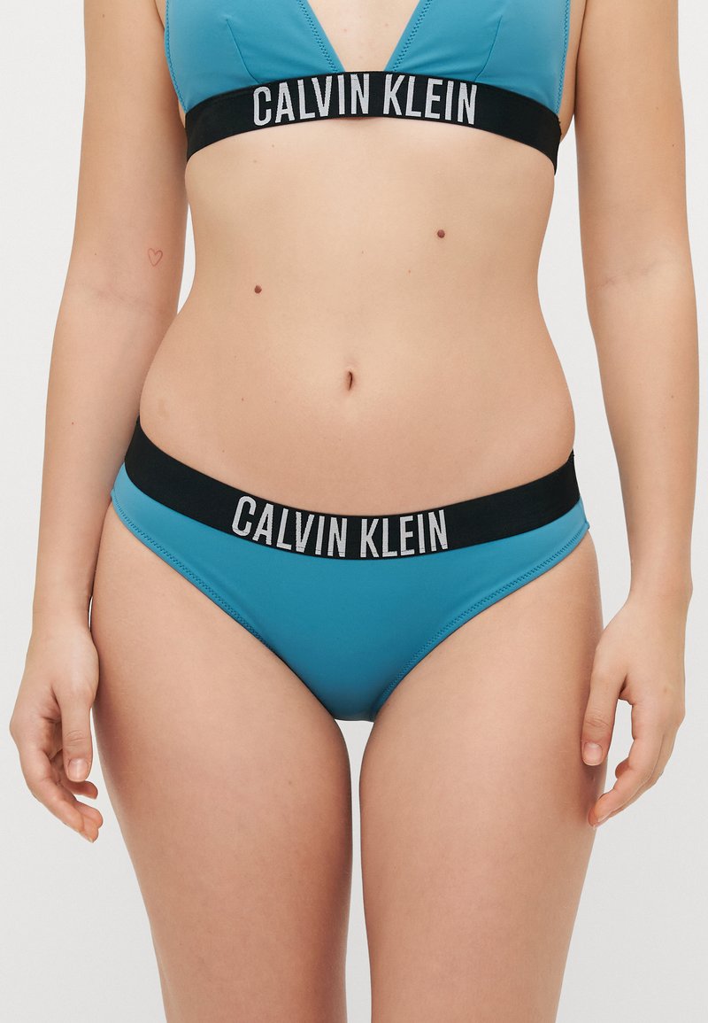 Top de bikini em triângulo turquesa com cintura preta com logotipo, apresentando uma textura suave e design minimalista. Letreiro da marca em branco na faixa.
