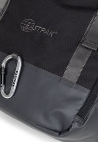 Černý látkový batoh s šedými akcenty, s texturovaným povrchem, logem a stříbrným karabinkovým klipem na boku.