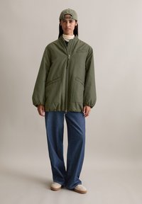 Olijfgroene gewatteerde jas met een ritssluiting aan de voorkant, grote zijzakken en elastische manchet, gecombineerd met losse denimbroeken en beige schoenen.