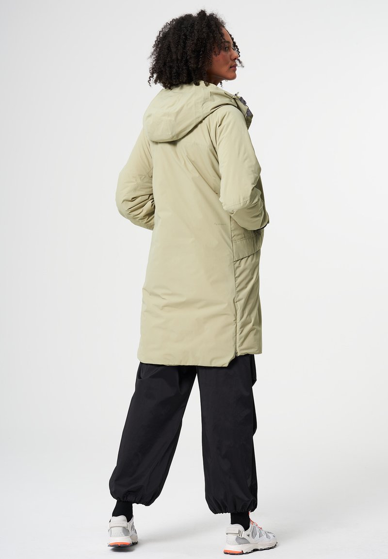 Lange, olivgrüne gepolsterte Jacke mit Kapuze, mit Reißverschluss und Seitentaschen, kombiniert mit lockeren schwarzen Hosen und Sneakers.