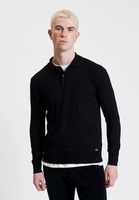 Zwart poloshirt met lange mouwen en kraag, voorzien van een drie-knoopsluiting. De stof lijkt glad, met subtiele stikdetails.