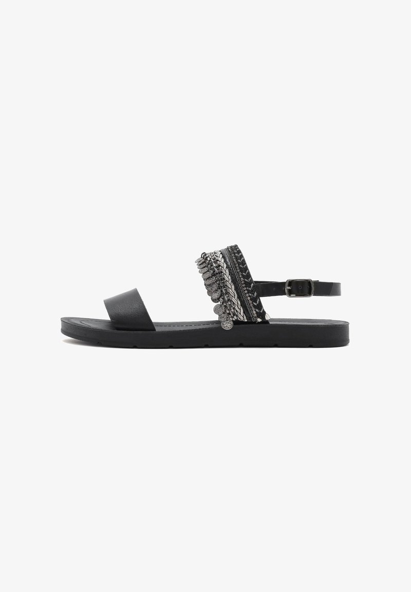 Sandal nera con suola piatta, ampia fascia frontale e una banda decorativa con accenti metallici e trecce. Fascetta regolabile alla caviglia con fibbia.