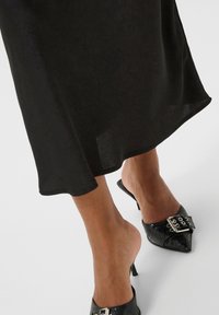 Jupe noire fluide avec une finition texturée, associée à des mules à talons noirs pointues avec une boucle décorative.