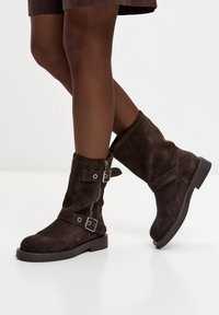 Ghete maro din piele întoarsă cu două catarame argintii și fermoar lateral. Talpă plată și design cu vârf rotunjit. Textură netedă.