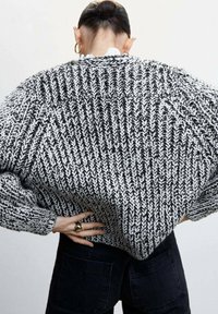 Knautschiger Strickpullover im Schwarz-Weiß-Fischgrätmuster. Übergrößenpassform mit gerippten Bündchen. Das Model trägt ihn mit schwarzen Hosen und zeigt eine lässige Silhouette.