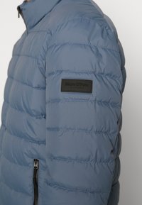 Giacca a vento blu con sezioni orizzontali, realizzata in tessuto liscio. Presenta una chiusura con zip e un piccolo etichetta nera sulla spalla con il nome del marchio.