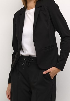 Persoon die een zwarte blazer met elleboogstukken draagt, een zwarte broek met trekkoord en een wit shirt, met één hand in de zak tegen een effen achtergrond.
