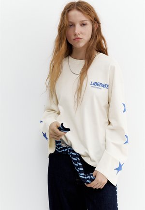 Ung kvinde med langt rødt hår iført en cremefarvet sweatshirt med blå stjerner og teksten "LIBERATE", der holder et stribet stofbælte.