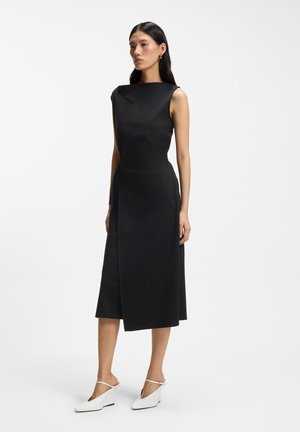 Woman with long dark hair wearing a sleeveless black midi dress and white pointed-heel shoes, standing against a plain light background.