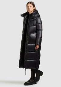 Lange zwarte pufferjas met een glanzende afwerking, oversized capuchon, voorzakken met rits en een aansluitende pasvorm, gedragen over zwarte schoeisel.