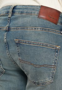 Κοντινό πλάνο πίσω τσέπης ανοιχτόχρωμου γαλάζιου τζιν με πορτοκαλί ραφές και καφέ δερμάτινο λογότυπο μάρκας Pepe Jeans London.