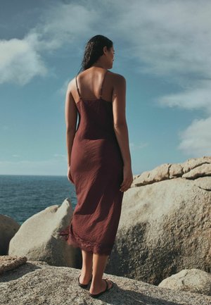 Naine pruunist varrukateta kleidis ja sandaalides seisab kivisel kaldal, vaadates osaliselt pilvise taeva all ookeani.