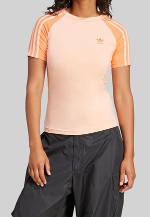 Kobieta nosząca dopasowaną bladoróżową koszulkę Adidas z pomarańczowymi rękawami i paskami, zestawioną z luźnymi czarnymi spodniami na jednolitym tle.