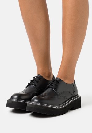 Chaussures en cuir noir à lacets avec des détails de brogue, semelle épaisse en plateforme noire et accents de couture blanche. Surfaces lisses et texturées visibles.