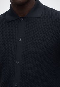 Mørk marineblå knappet strikkeskjorte med teksturert vaffelmønster båret av en person, som viser krage og øvre del av overkroppen.