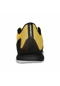 Zapatilla de tenis negra y amarilla con parte superior de malla, diseño de talón redondeado y logotipo prominente en el talón. Suela blanca con patrón texturizado.