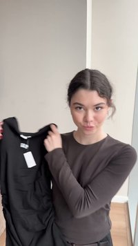 Tânără cu păr închis la culoare ține un tricou negru cu mâneci lungi, cu o etichetă albă, într-un spațiu interior luminos și minimalist.