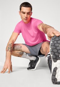 Rosa Sportshirt, graue Shorts, schwarze Laufschuhe mit weißen Sohlen, Tattoos an den Beinen, dehnende Pose vor einem neutralen Hintergrund.