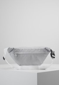 Sac banane gris en tissu léger, doté d'une poche zippée à l'arrière, d'une sangle blanche ajustable et d'un design minimaliste sans motifs.