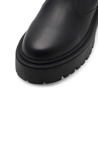Czarny skórzany botek za kostkę z zaokrąglonym noskiem, grubą, teksturowaną gumową podeszwą oraz ozdobnym przeszyciem wzdłuż krawędzi buta.