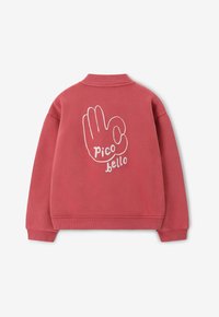 Sweatshirt rouge avec un col côtelé. Présente une illustration de main blanche et le texte "pico bello" au dos. Texture douce, design décontracté.