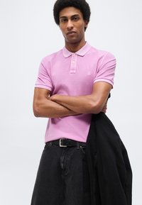 Άνδρας με σταυρωμένα χέρια, φορώντας ροζ πόλο μπλούζα και μαύρα τζιν, κρατώντας μαύρο σακάκι, μπροστά σε απλό λευκό φόντο.