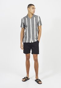 Koszula w czarno-białe wzory z krótkim rękawem i kołnierzykiem, zestawiona z czarnymi szortami i czarnymi sandałami, na białym tle.