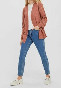 Svetr pletený rezavé barvy s otevřeným předním dílem, doplněný světle modrými džíny s vysokým pasem a bílými botami; uvolněný střih.