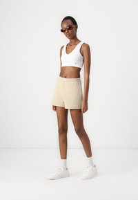 Bijela v-neck crop majica, bež kratke hlače s elastičnim pojasom i bijele tenisice. Model nosi crvene sunčane naočale i stoji na jednoličnoj pozadini.