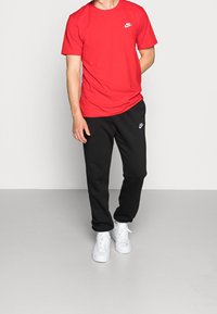 Muž v červeném tričku Nike, černých teplácích Nike a bílých teniskách stojí na světlém jednobarevném pozadí.