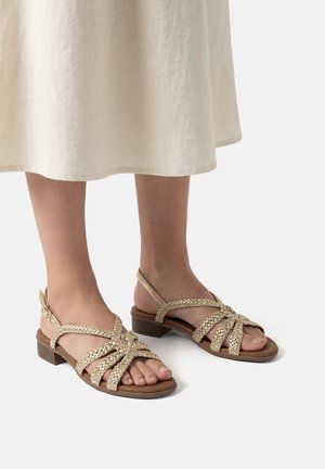 Piedi che indossano sandali beige con cinturini intrecciati e tacchi bassi marroni, abbinati a una gonna color crema che scende sotto le ginocchia.