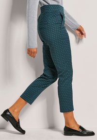 Türkis gemusterte Hose mit hoher Taille, Kapuzenschnitt und geometrischem Design, kombiniert mit glänzenden schwarzen Loafers.