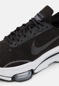 Černé semišové tenisky s perforovaným designem, šedými akcenty a viditelnou vzduchovou komořen u paty. Disponují texturovanými tkaničkami a logem.