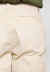 Primer plano de pantalones beige con bolsillo trasero de ribete y detalles de costura, combinados con una blusa de color verde oliva.
