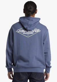 Modrá mikina s kapucňou a veľkým grafickým logom na zadnej strane, obsahujúca biele a zelené písmo. Vyrobená z mäkkej a odolnej látky.