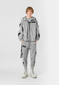 Persona joven de pie, vistiendo una sudadera con capucha gris claro y pantalones deportivos con parches de alas en blanco y negro, y botas blancas con cordones, con las manos en los bolsillos.