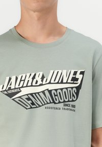 Mintgrøn bomulds T-shirt med fed sort og hvid grafisk tekst: "JACK & JONES," "DENIM GOODS," med rund hals og korte ærmer.