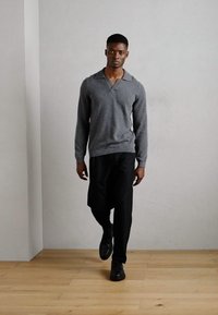 Gray, knitted sweater with a v-neck design, paired with black trousers and black shoes. Standing on wooden flooring against a plain wall.