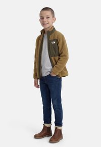 Garçon portant une veste polaire marron et olive, un t-shirt gris, un jean bleu et des bottes marron, debout avec une main dans la poche et souriant.