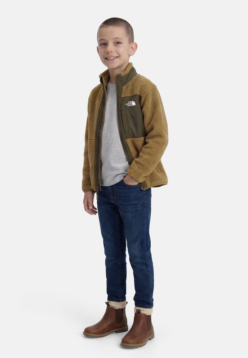 Garçon portant une veste polaire marron et olive, un t-shirt gris, un jean bleu et des bottes marron, debout avec une main dans la poche et souriant.